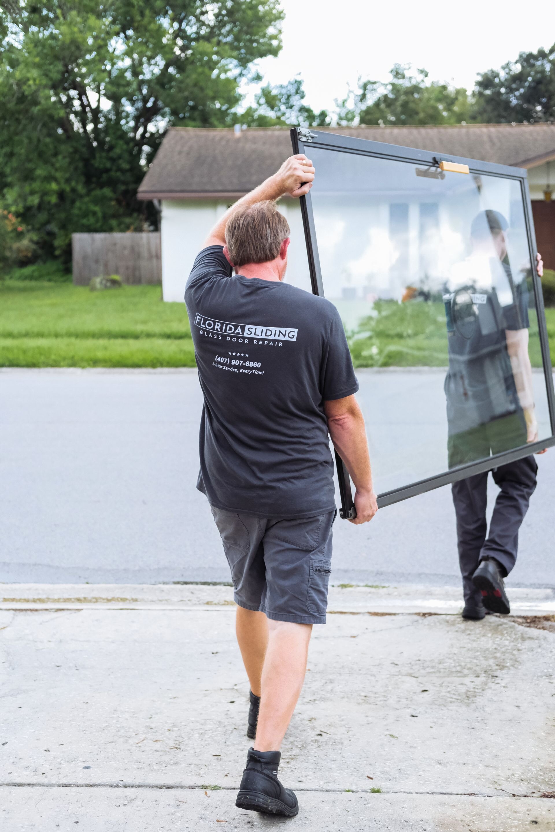 Two Florida Sliding Glass Door Repair technicians in branded shirts carrying a sliding glass door at a Central Florida home